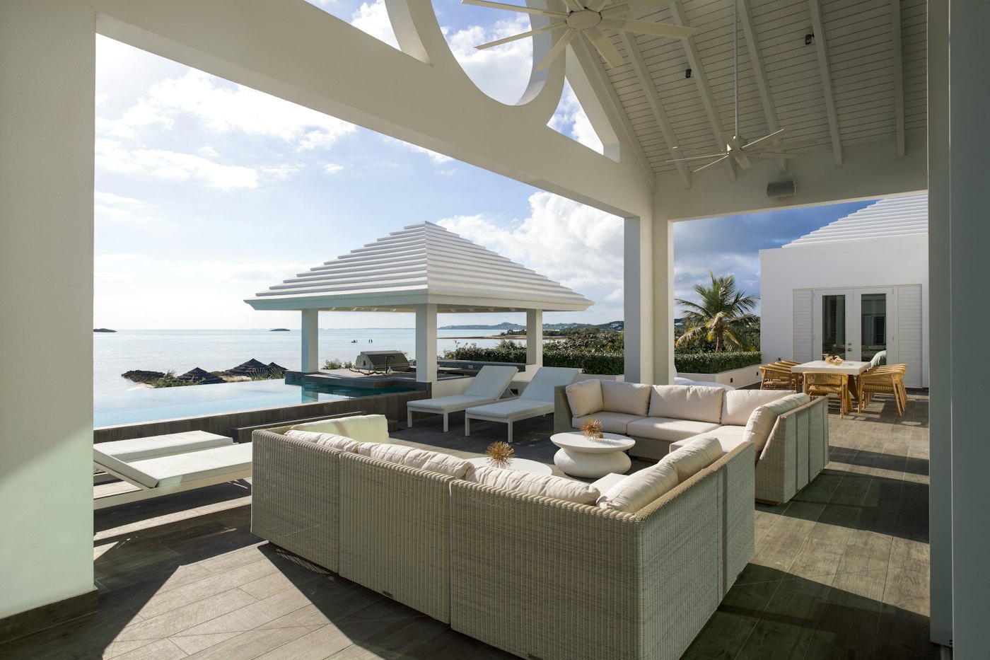 A shaded villa terrace with white sofas beside an infinity pool overlooking the ocean